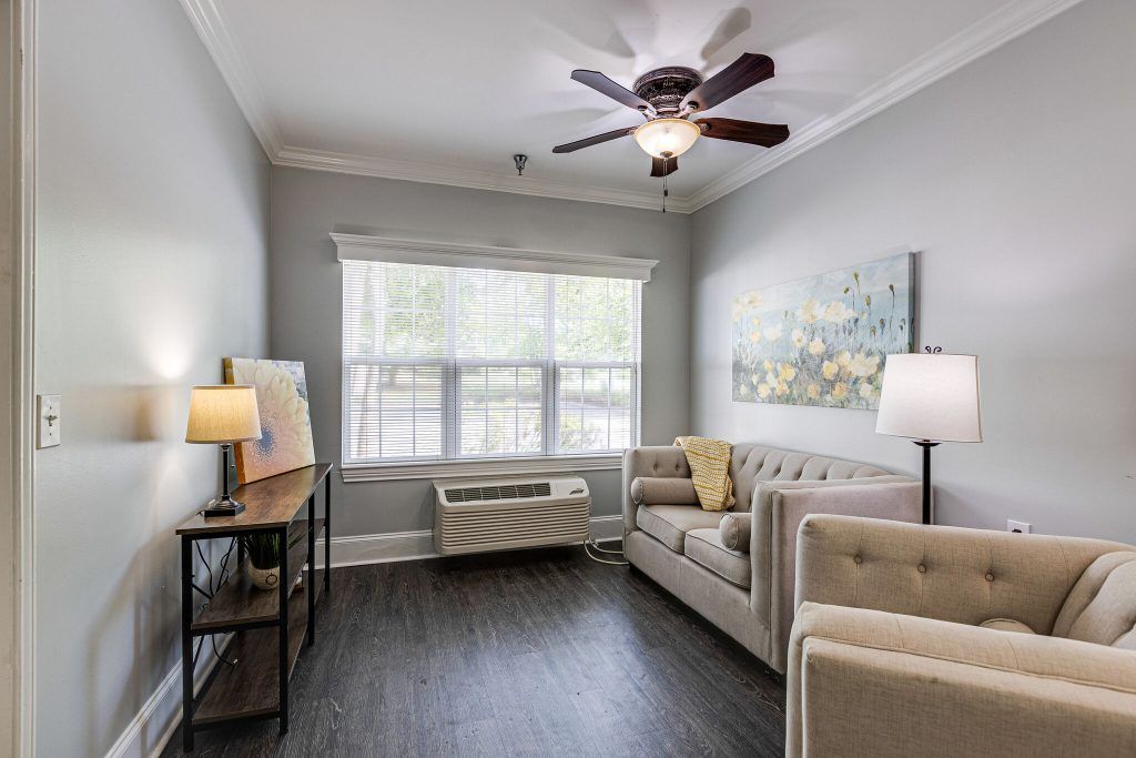 A modern living room with a comfortable sofa, ceiling fan, and decorative lamps, featuring large windows and stylish artwork.