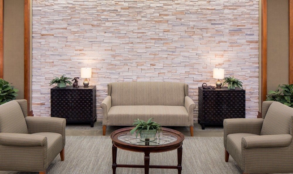 A cozy living room with a beige sofa, armchairs, a round wooden coffee table, and decorative lamps against a brick wall.