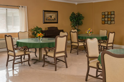 A welcoming dining room setup featuring several green-topped tables surrounded by beige chairs, enhanced by decorative plants and a floral centerpiece.