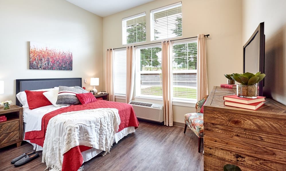 A bright and modern bedroom with large windows, wood flooring, and red accent decor.