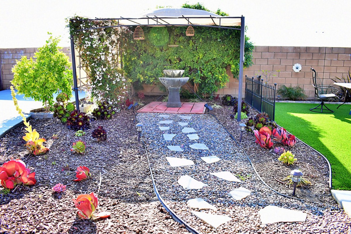A garden with a stone pathway leading to a pergola and fountain, surrounded by gravel and plants.