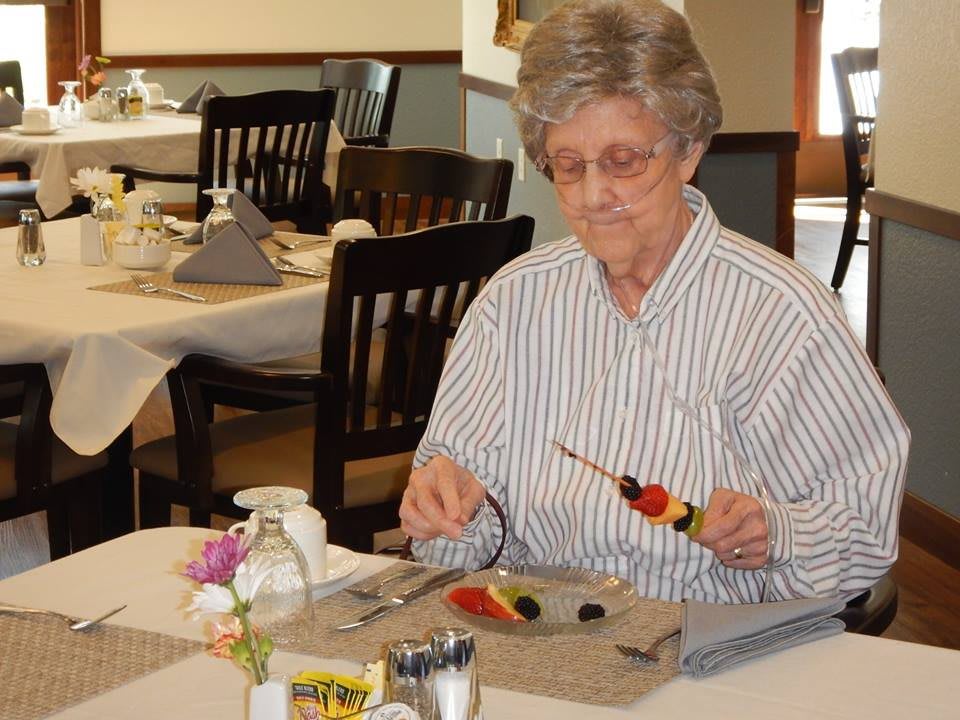 An elderly woman prepares a fruit skewer at the dining table.