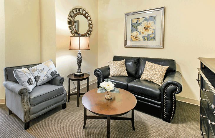 A cozy living room with a black leather sofa, an armchair, and a round wooden coffee table, complemented by a lamp, mirror, and floral painting on the wall.