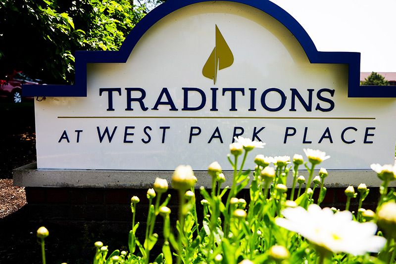 A sign for Traditions at West Park Place amidst greenery and flowers.