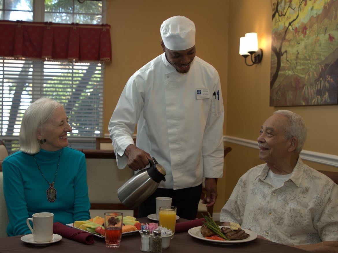 A chef serving two seniors at a dining table.
