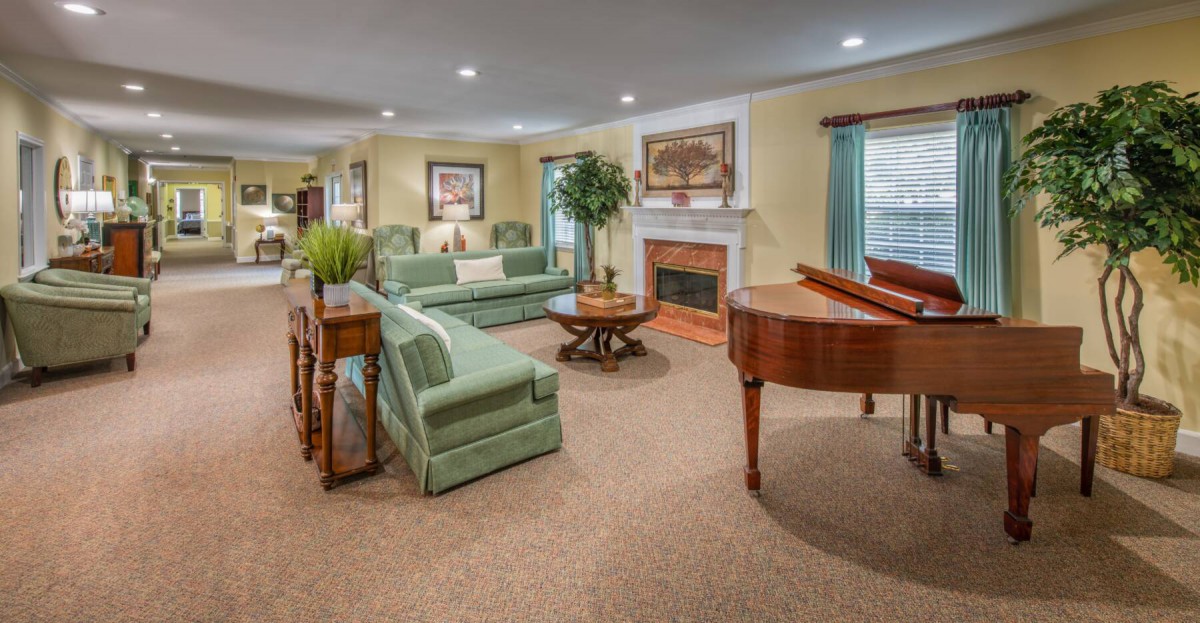 A spacious living room featuring a grand piano, cozy sofas, and a fireplace, elegantly decorated with artwork and greenery.