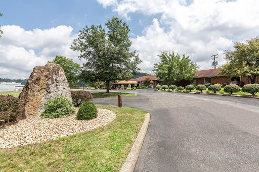 A scenic view of a landscaped entrance to a building along a winding road, surrounded by trees and clouds.