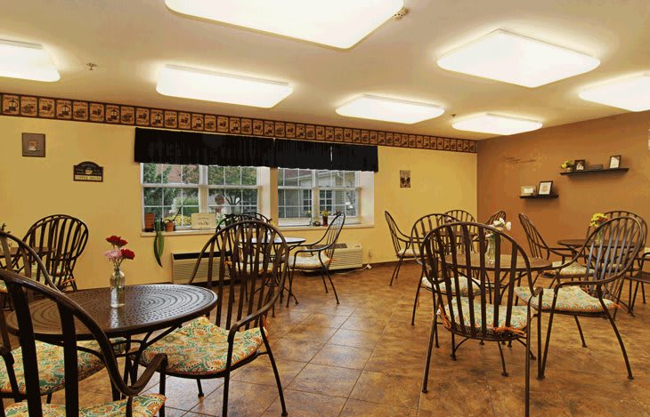 A cozy dining room with multiple tables and chairs, featuring large windows and a wooden floor.