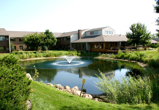 A serene landscape featuring a tranquil pond with a fountain, surrounded by lush greenery and a modern building.
