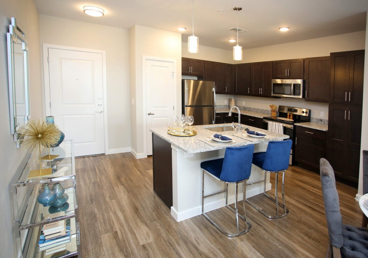 A modern kitchen with stainless steel appliances, featuring an island counter with blue bar stools and hardwood flooring.