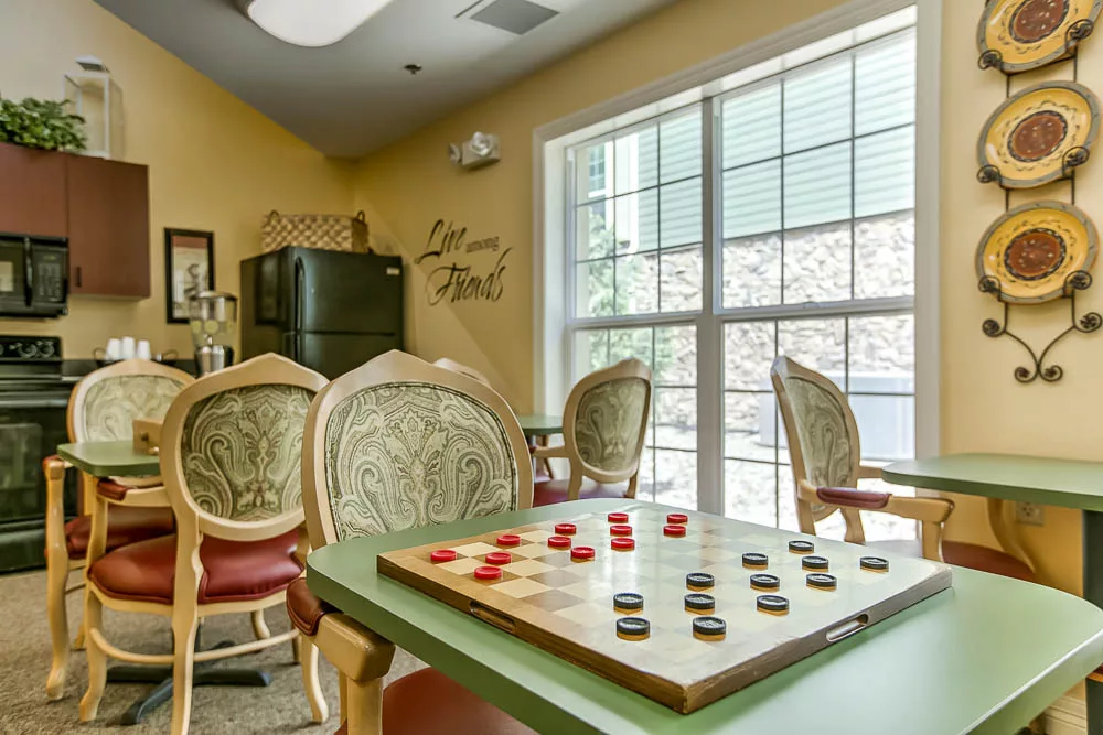 A cozy game room featuring a checkers board set up on a table, surrounded by chairs and a window letting in natural light.