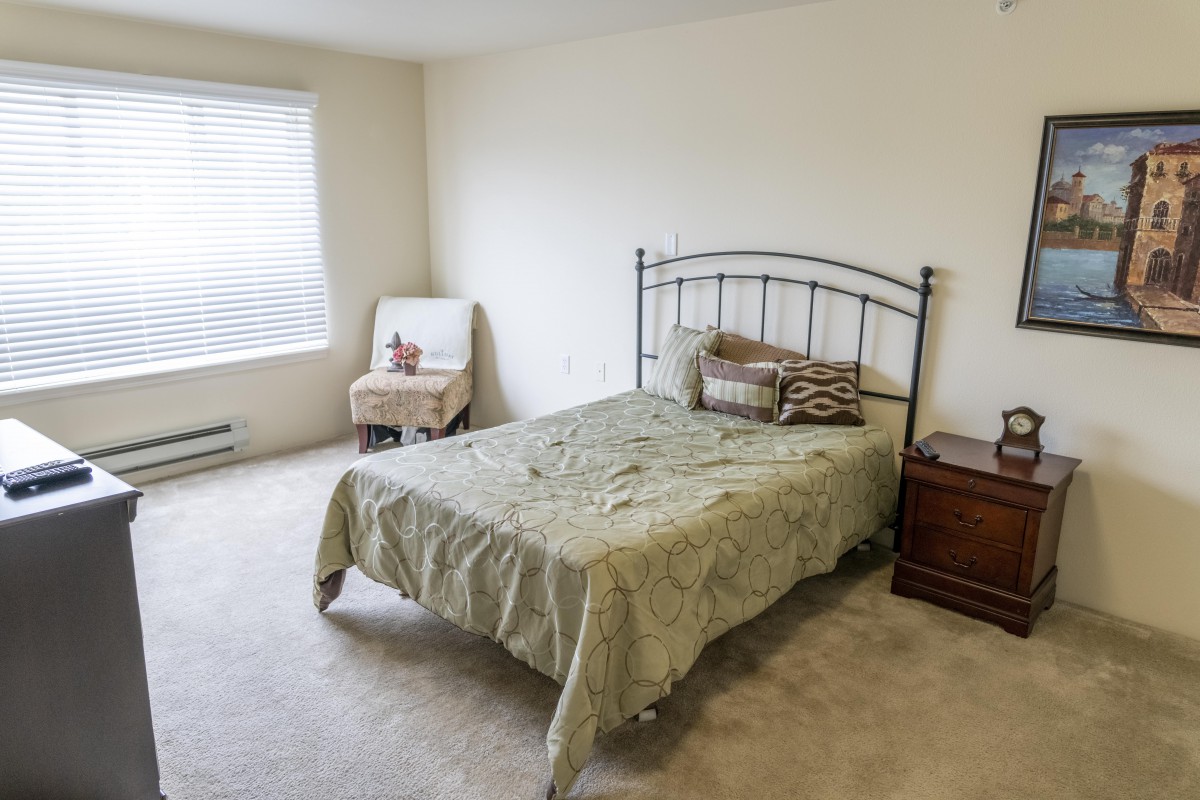A simple bedroom with a metal-framed bed, a small chair by the window, and a wooden nightstand.