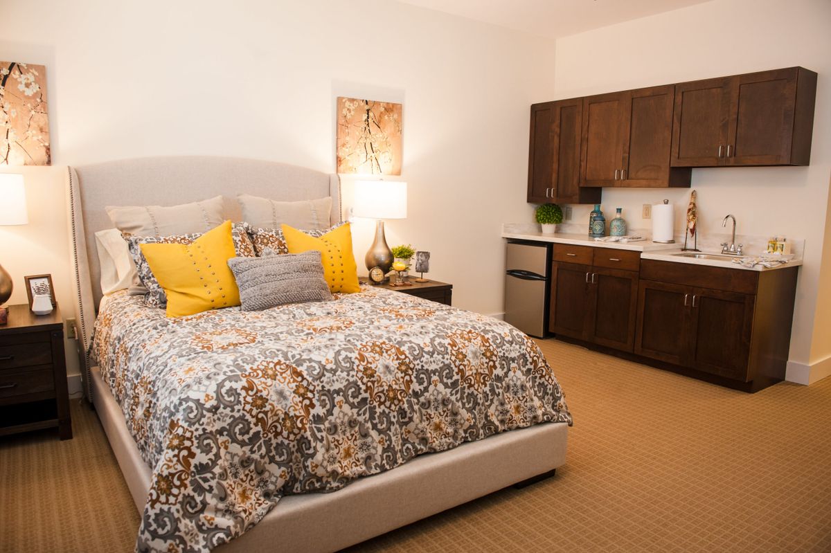 A cozy bedroom with a plush bed adorned with patterned bedding and vibrant pillows, complemented by a kitchenette in the corner.