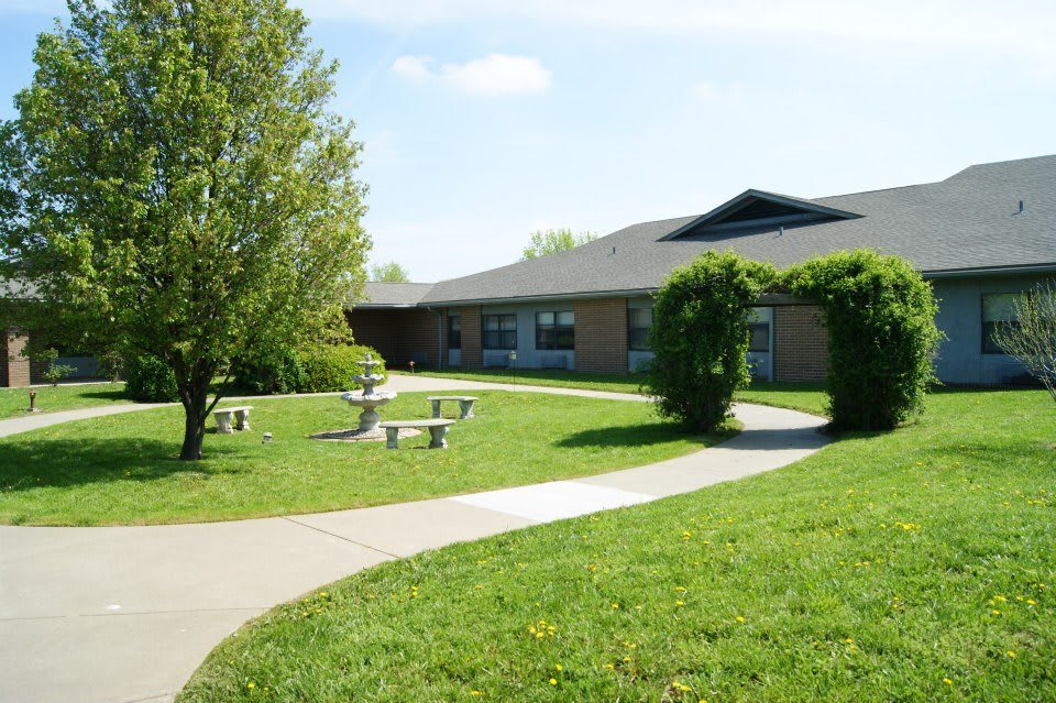 A tranquil outdoor area with a pathway leading to a school building, surrounded by greenery and seating benches.