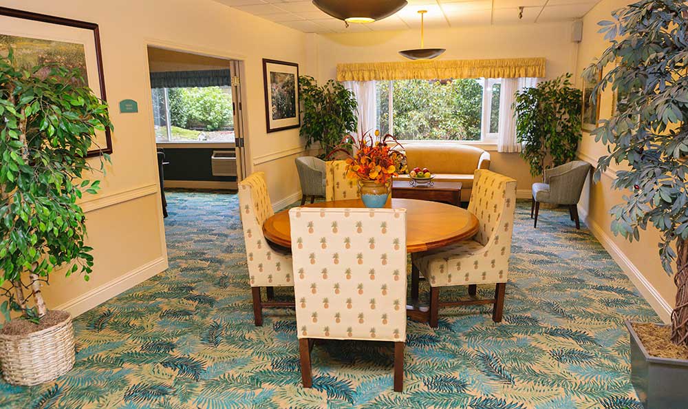 A cozy dining room with a round table and upholstered chairs, featuring a patterned carpet and large windows allowing natural light.