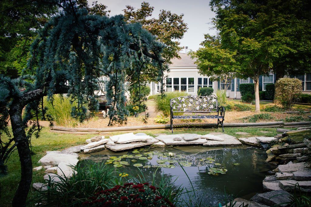 A tranquil garden with a pond, surrounded by lush greenery and a quaint building in the background.