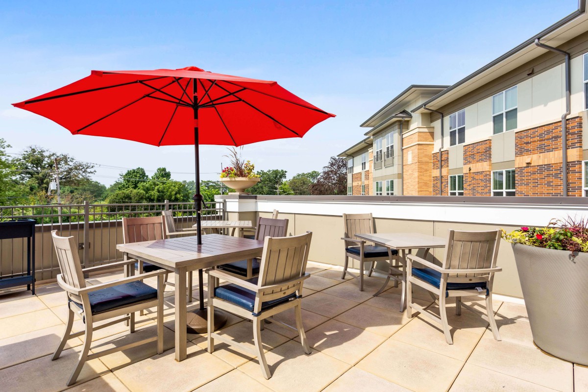 A sunny rooftop patio with tables, chairs, and a red umbrella.