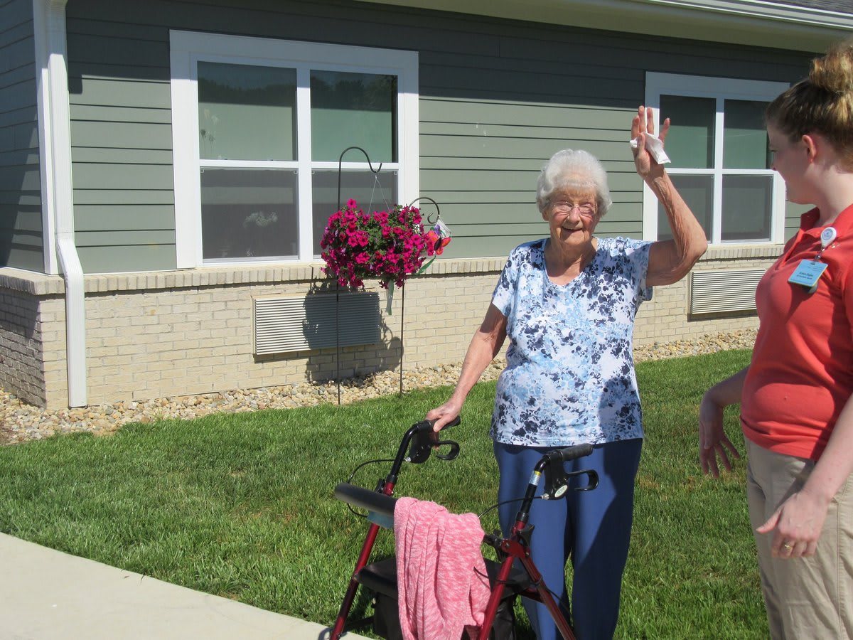 An elderly woman waves cheerfully while standing outside with her walker on a sunny day.