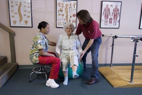 An elderly person receiving assistance with physical therapy exercises from two caregivers in a healthcare setting.