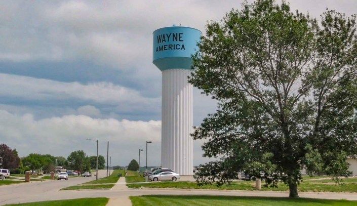 A water tower labeled 'Wayne America' stands tall beside a road and trees under a cloudy sky.