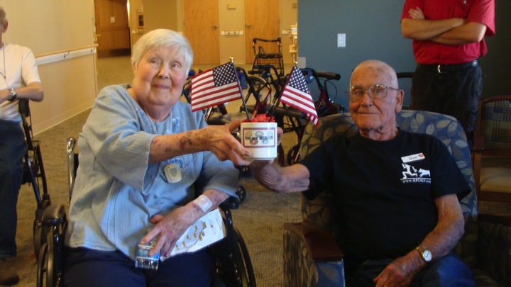 An elderly couple enjoying a moment together, holding a coffee mug and surrounded by American flags in an indoor setting.