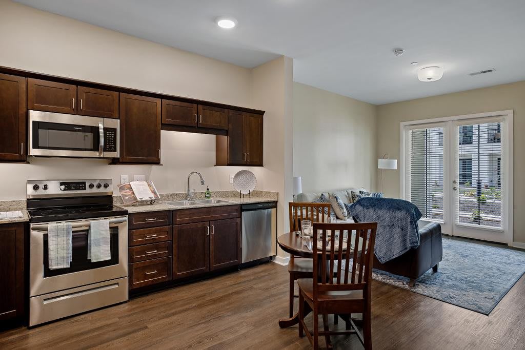 A modern apartment interior featuring a kitchen with dark cabinets, a small dining table, and an adjacent living area.