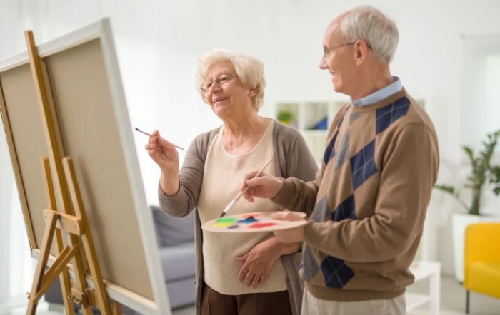 An elderly couple painting together at an easel.