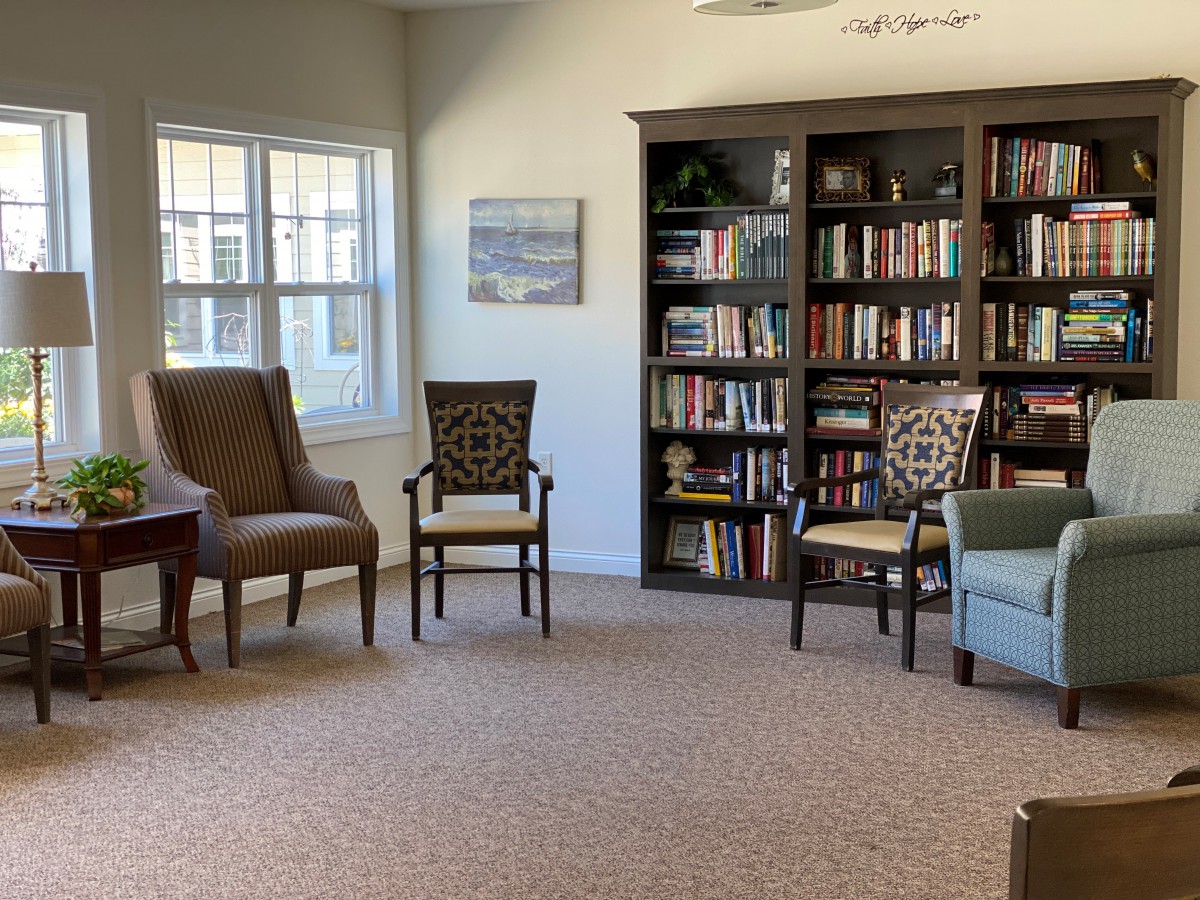 A cozy reading room with bookshelves, various chairs, and natural light from a window.