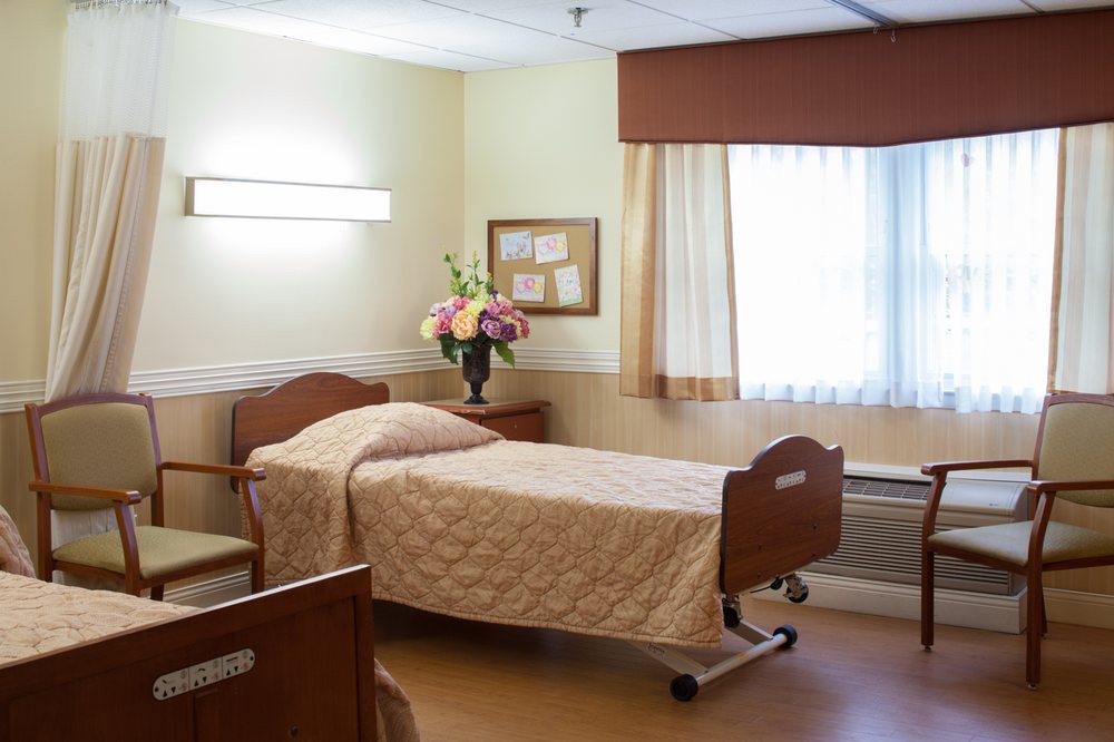 A well-kept hospital room with a single patient bed, decorated with flowers and natural light coming through the window.