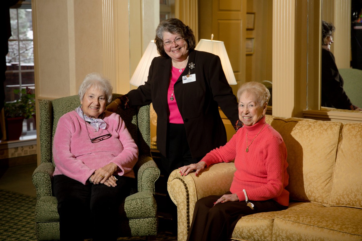 Three women, two seated and one standing, smiling indoors.