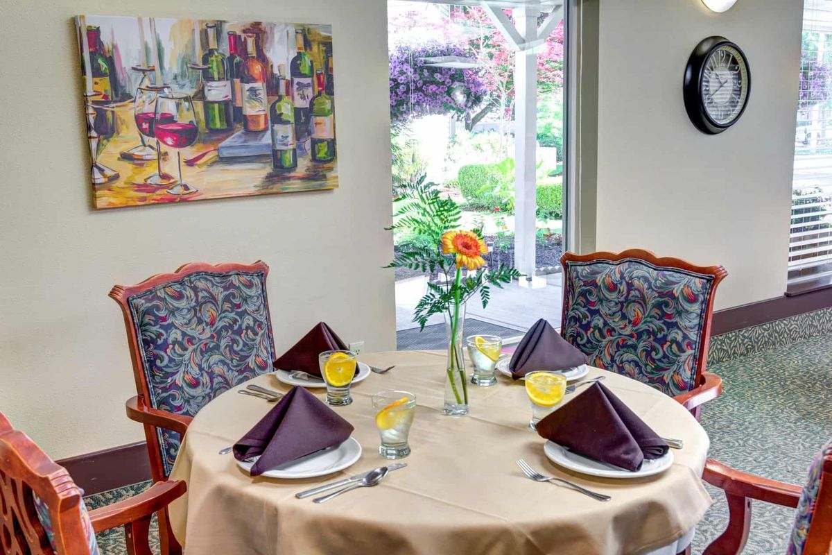 A neatly set dining table with purple napkins and a flower vase, next to a large window with a garden view.