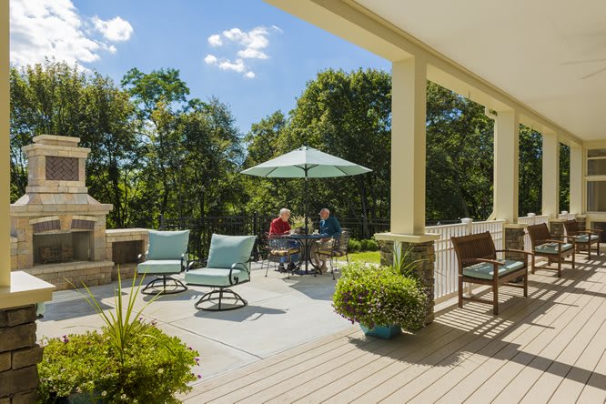 A sunny outdoor patio with people sitting under an umbrella, surrounded by lush greenery.