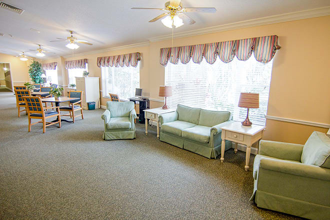 A cozy living room with sofas and armchairs arranged around tables, featuring large windows with curtains.