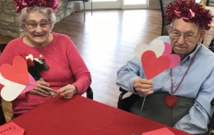 Two elderly women sitting at a table, wearing festive hats and holding paper hearts during a celebration.