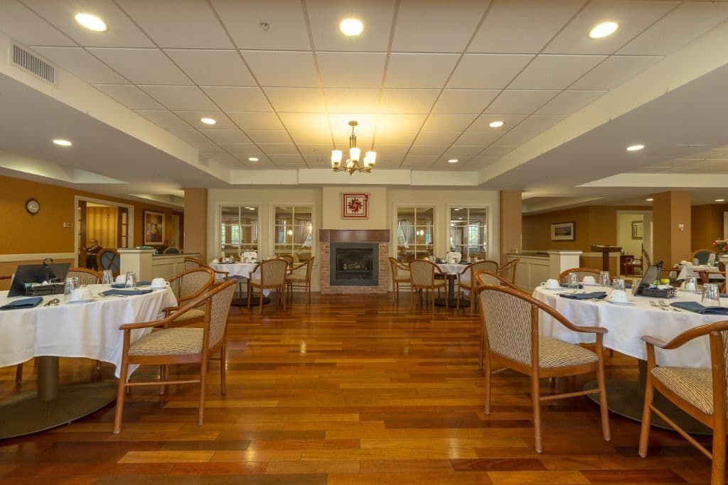 A cozy dining area with neatly arranged tables and chairs, featuring a wooden floor and a central chandelier.