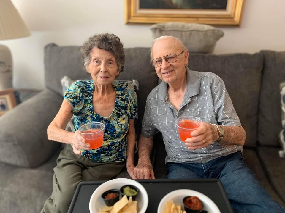 An elderly couple sitting on a sofa, enjoying drinks and snacks.