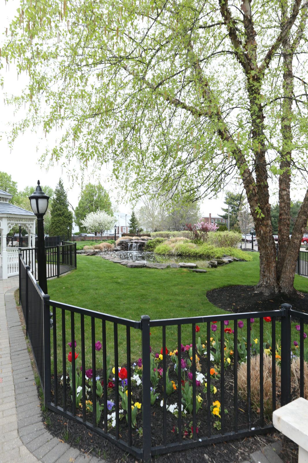 A picturesque garden scene with a black fence, lush green lawn, colorful flowers, and large trees.