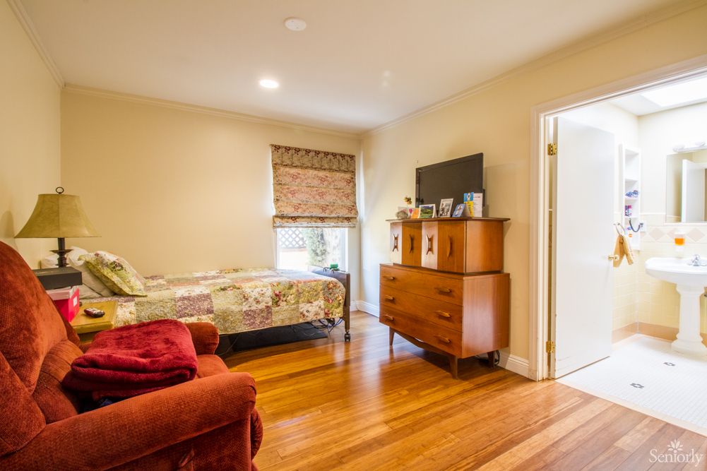 A cozy bedroom with hardwood flooring, featuring a single bed, wooden dresser with a TV on top, and an armchair near a window with blinds.