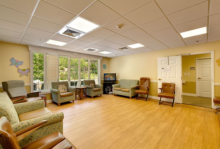 A cozy living room with comfortable chairs, a sofa, and a television, featuring large windows and hardwood flooring.