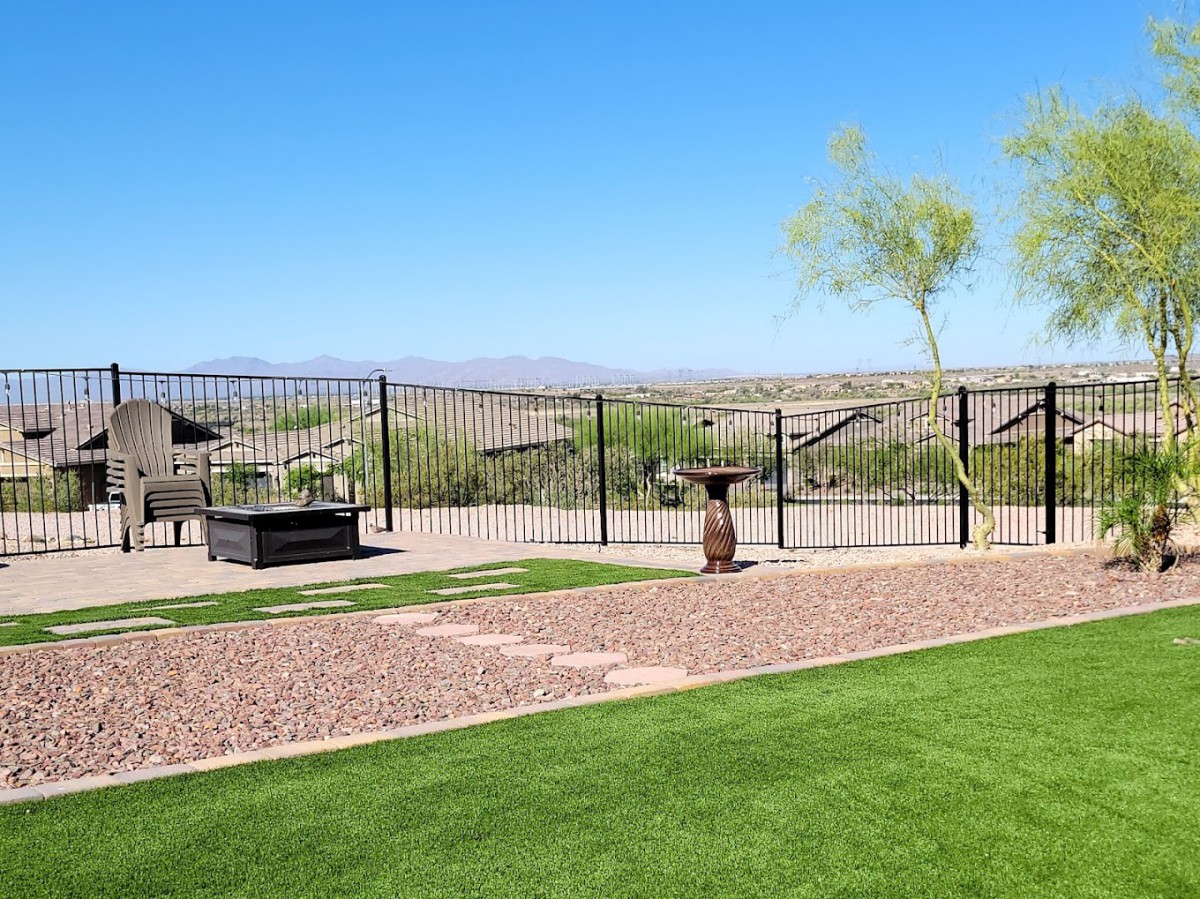 A backyard with a well-maintained lawn, small patio area, and desert landscape beyond a metal fence.