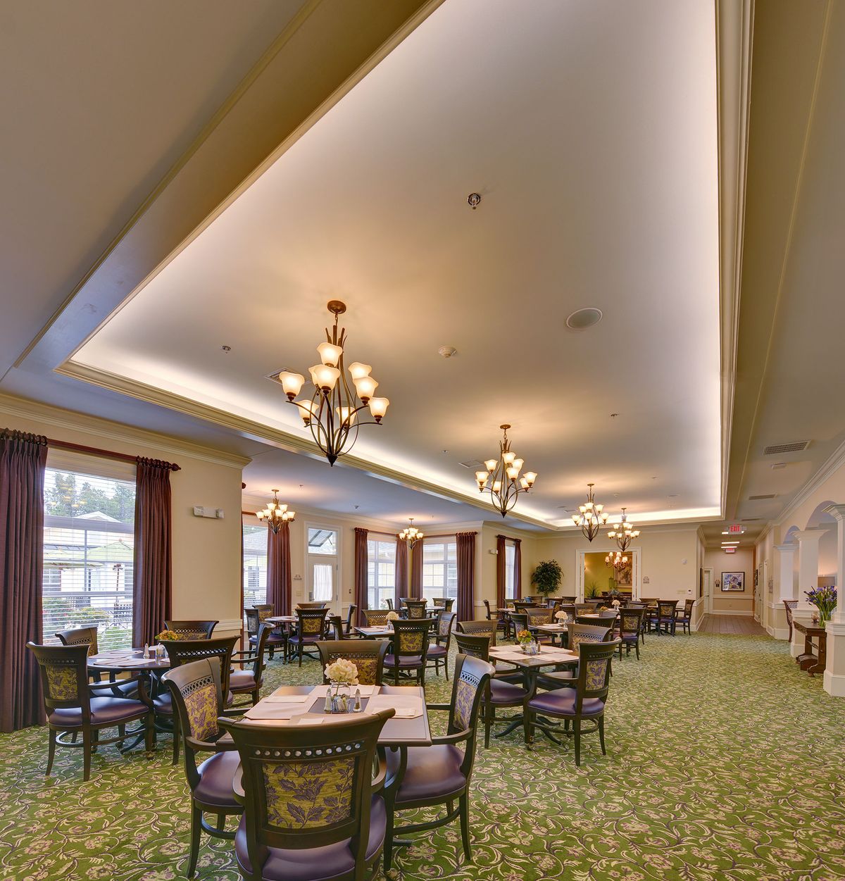 An elegant dining room with chandeliers, tables, and chairs, featuring a carpeted floor and large windows.