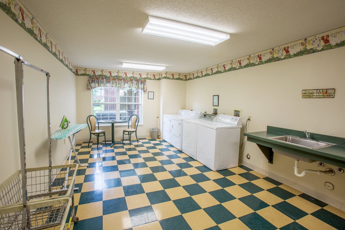 A well-lit laundry room with checkered flooring, featuring washing machines and a small seating area by the window.