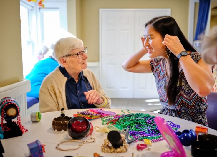 An elderly woman and a younger woman enjoy crafting together, surrounded by colorful beads on a table.