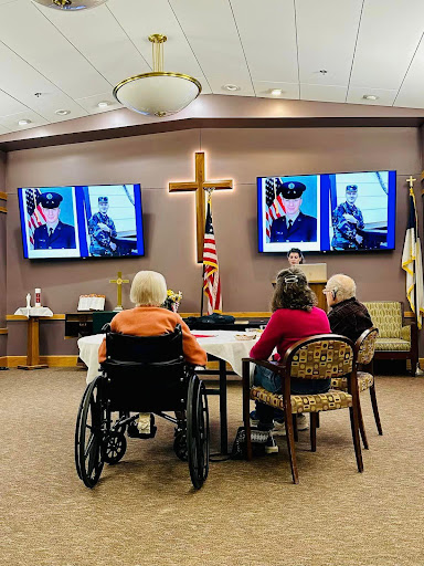 A gathering of senior citizens in a community center paying tribute to veterans, featuring images displayed on television screens.