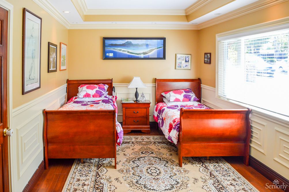 A cozy bedroom featuring two wooden twin beds with floral bedding and a central nightstand, adorned with paintings and a patterned carpet.