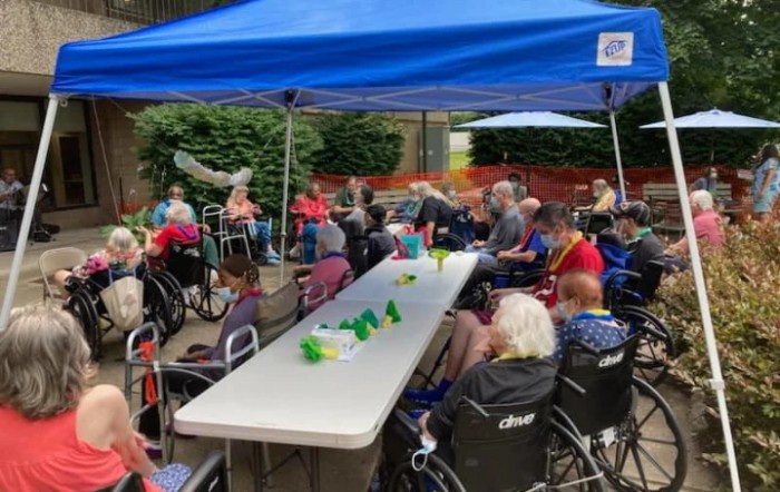 A community gathering of elderly people in wheelchairs under a blue tent outdoors.