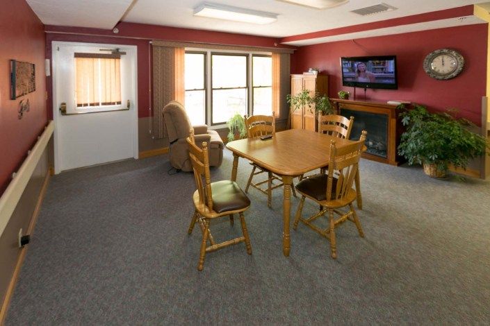 A cozy living room with a wooden dining table and chairs, a recliner, television, and indoor plants against red walls.