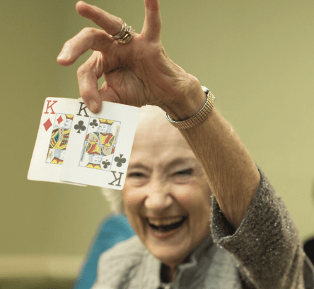 An elderly woman joyfully holding up a pair of playing cards.