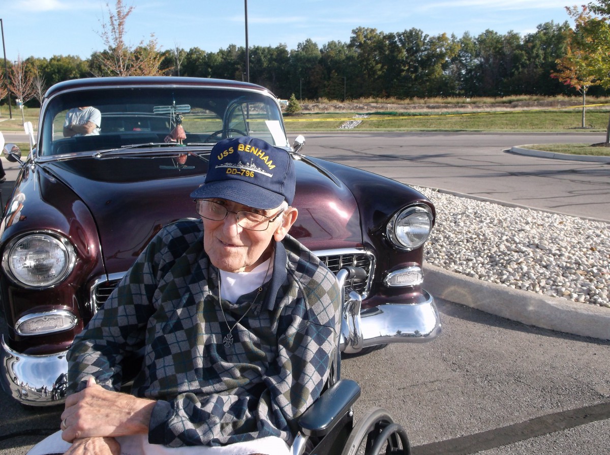 An elderly man in a wheelchair sits in front of a vintage car on a sunny day.