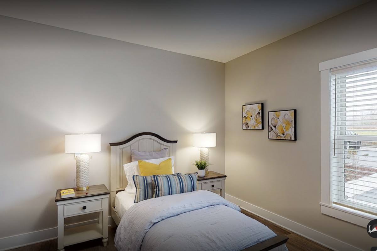 A cozy bedroom featuring a single bed with decorative pillows, flanked by two nightstands with lamps. The room is adorned with wall art for an added touch of style.
